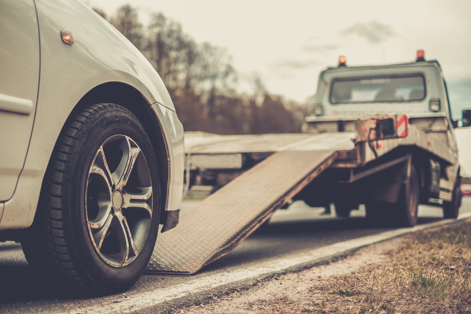 A white car positioned to drive onto the metal ramp of a parked tow truck on the side of a road.