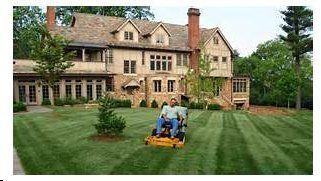 A person mows a large, neatly striped lawn in front of a multi-story stone home.