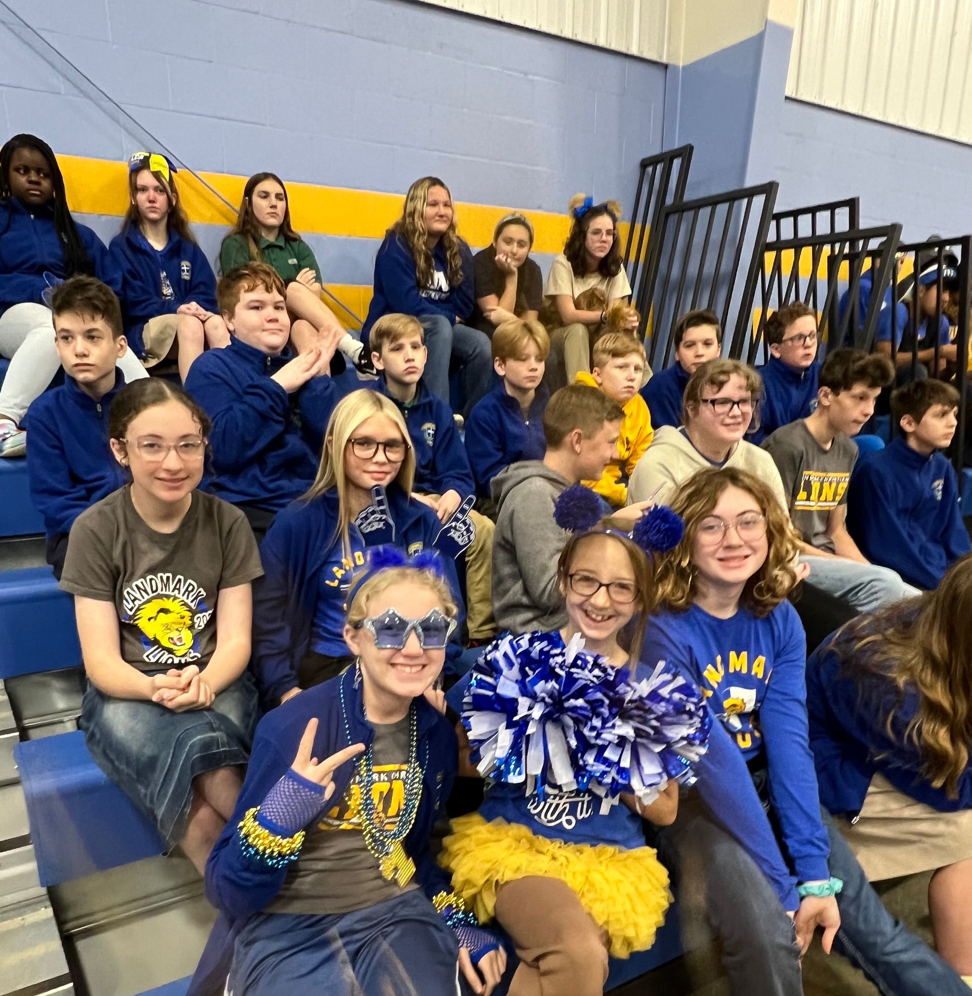 Students in blue and gold spirit wear sit on bleachers, smiling and posing with pom-poms.