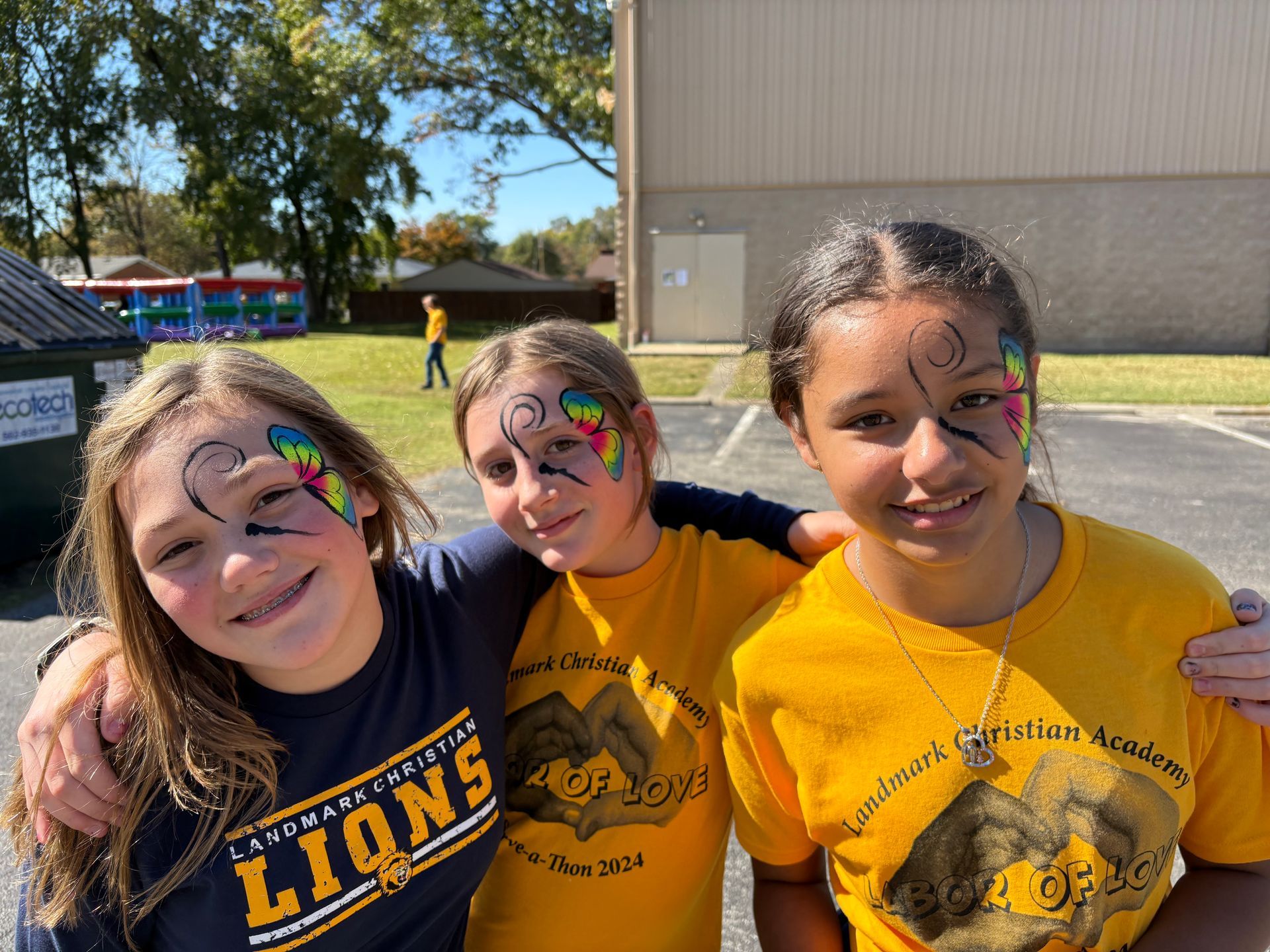 Three smiling kids in yellow shirts with face paint standing outdoors on a sunny day