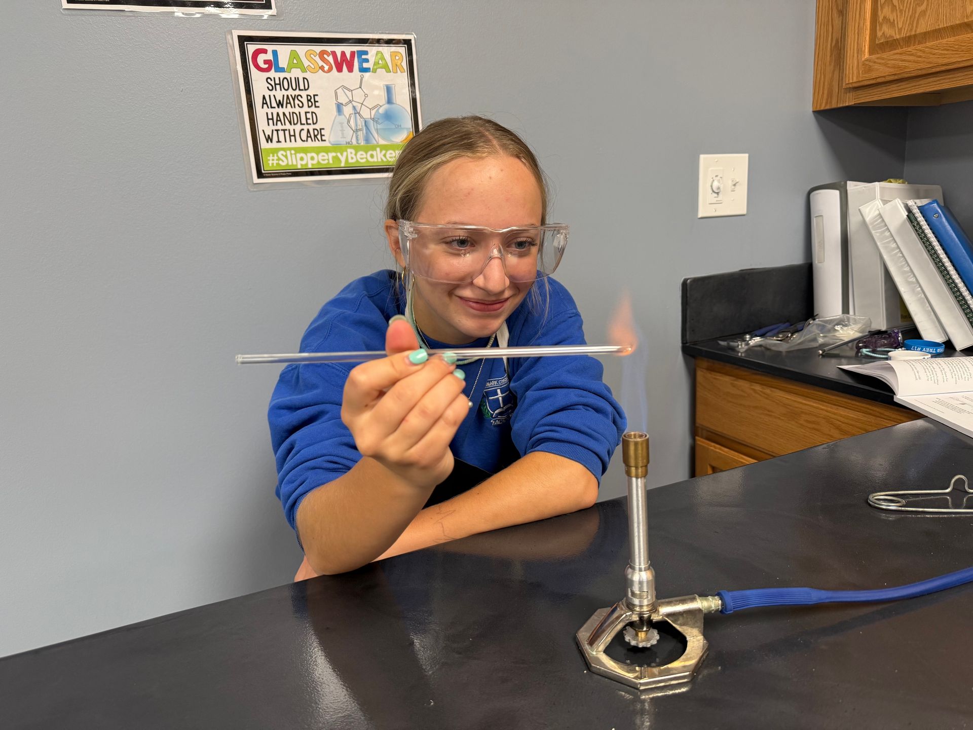 Student in safety goggles heating a thin rod over a lab Bunsen burner in a classroom.