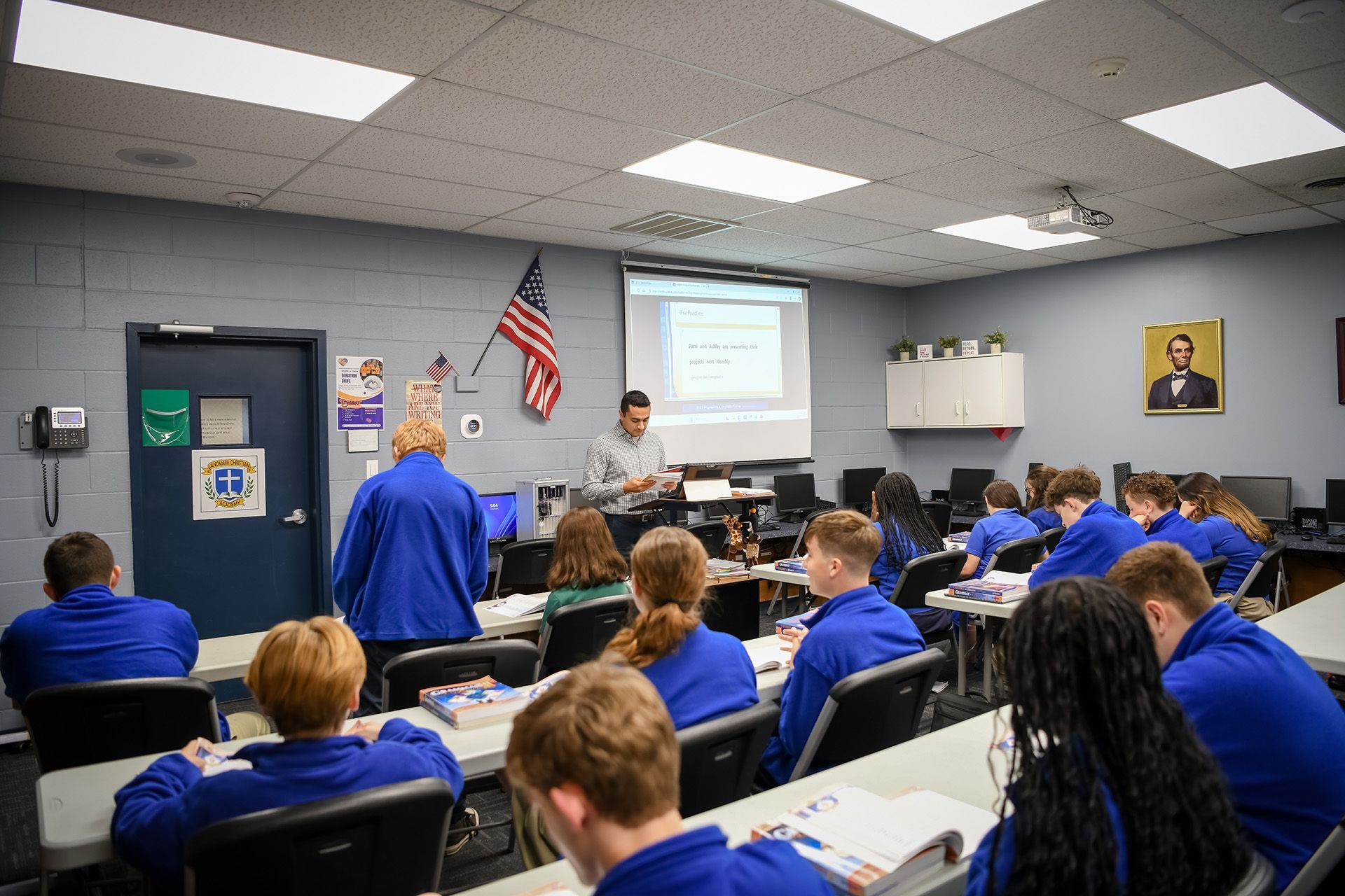 Classroom presentation with students in blue uniforms facing a teacher and projected slides on the wall