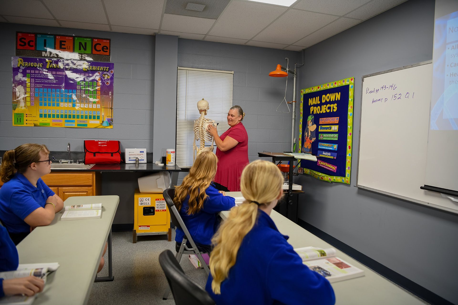 Teacher demonstrating on skeleton model in a classroom with students seated at desks