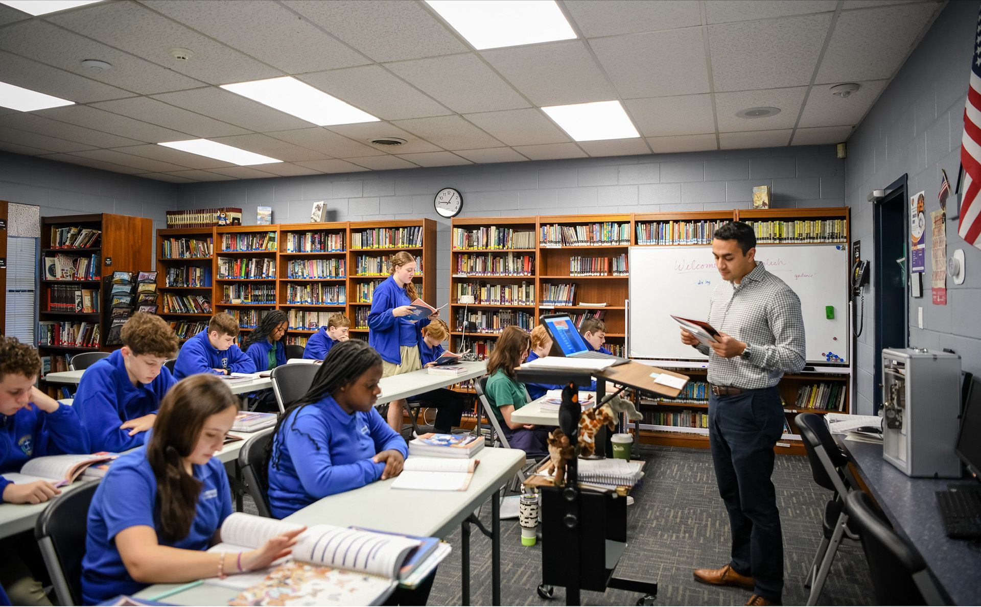 Students in a classroom reading while a teacher stands at the front holding papers