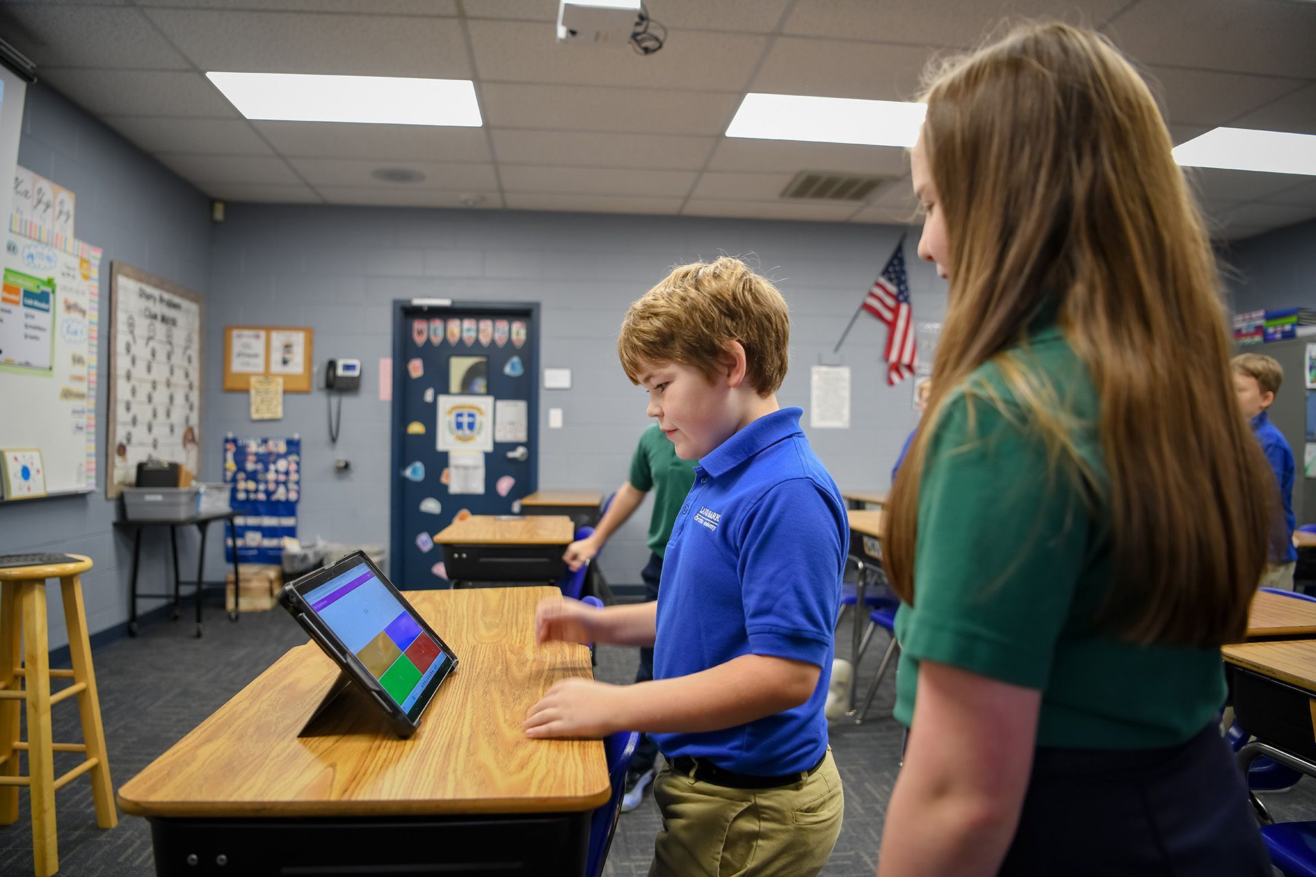 Students using a tablet at a classroom table while a teacher stands nearby.