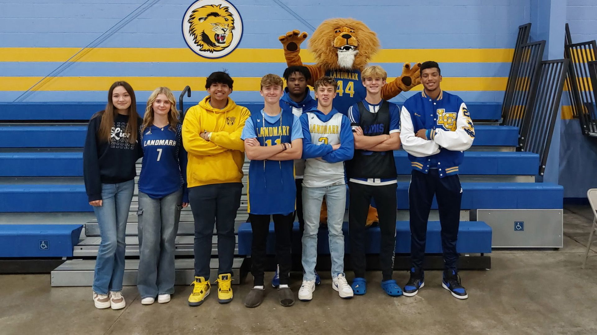 Seven students pose in front of blue bleachers with a lion mascot in a gymnasium.