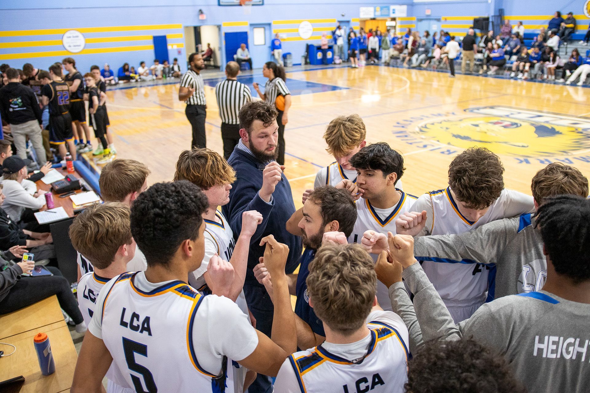Basketball team huddles with coach on the sideline during a game in a bright indoor gym.
