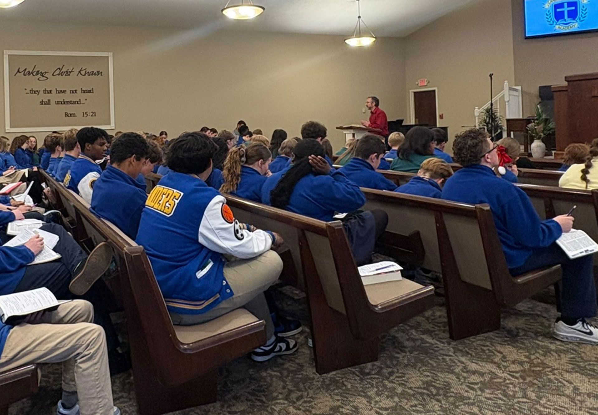 Audience in blue jackets seated in church pews, listening to a speaker at the front.