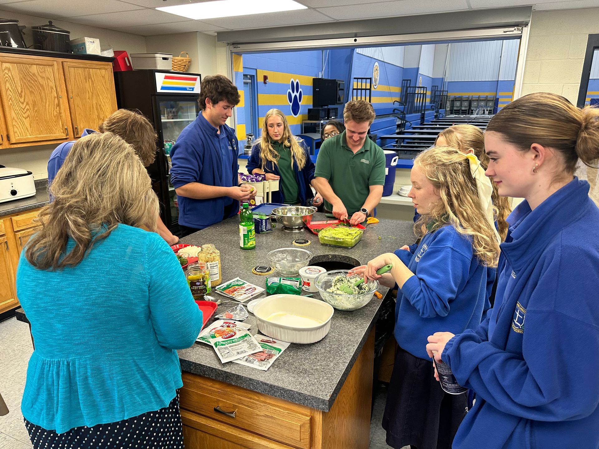 Students gather around a kitchen island preparing food in a bright classroom kitchen.