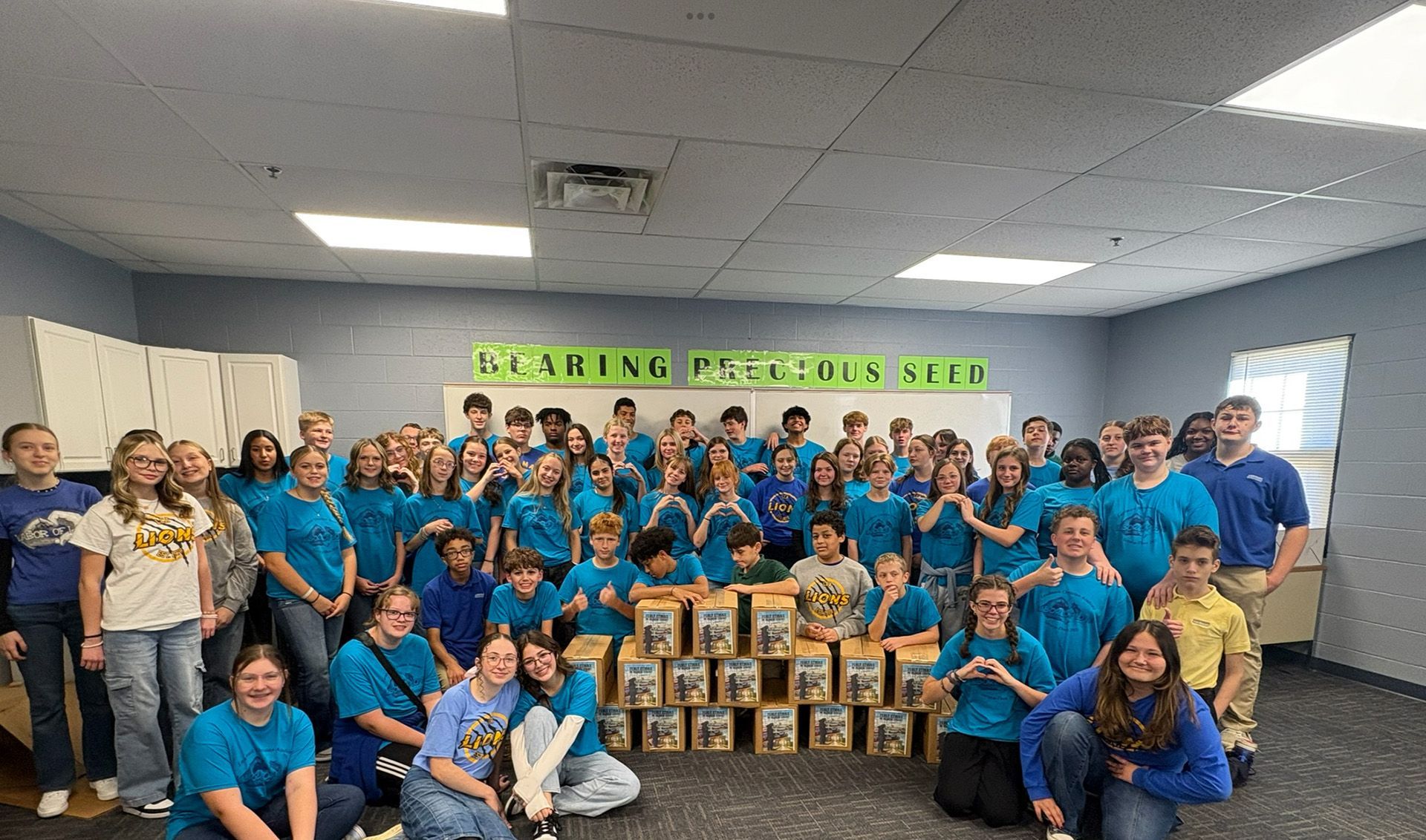 Large group in blue shirts posing indoors with stacked boxes and a green banner overhead
