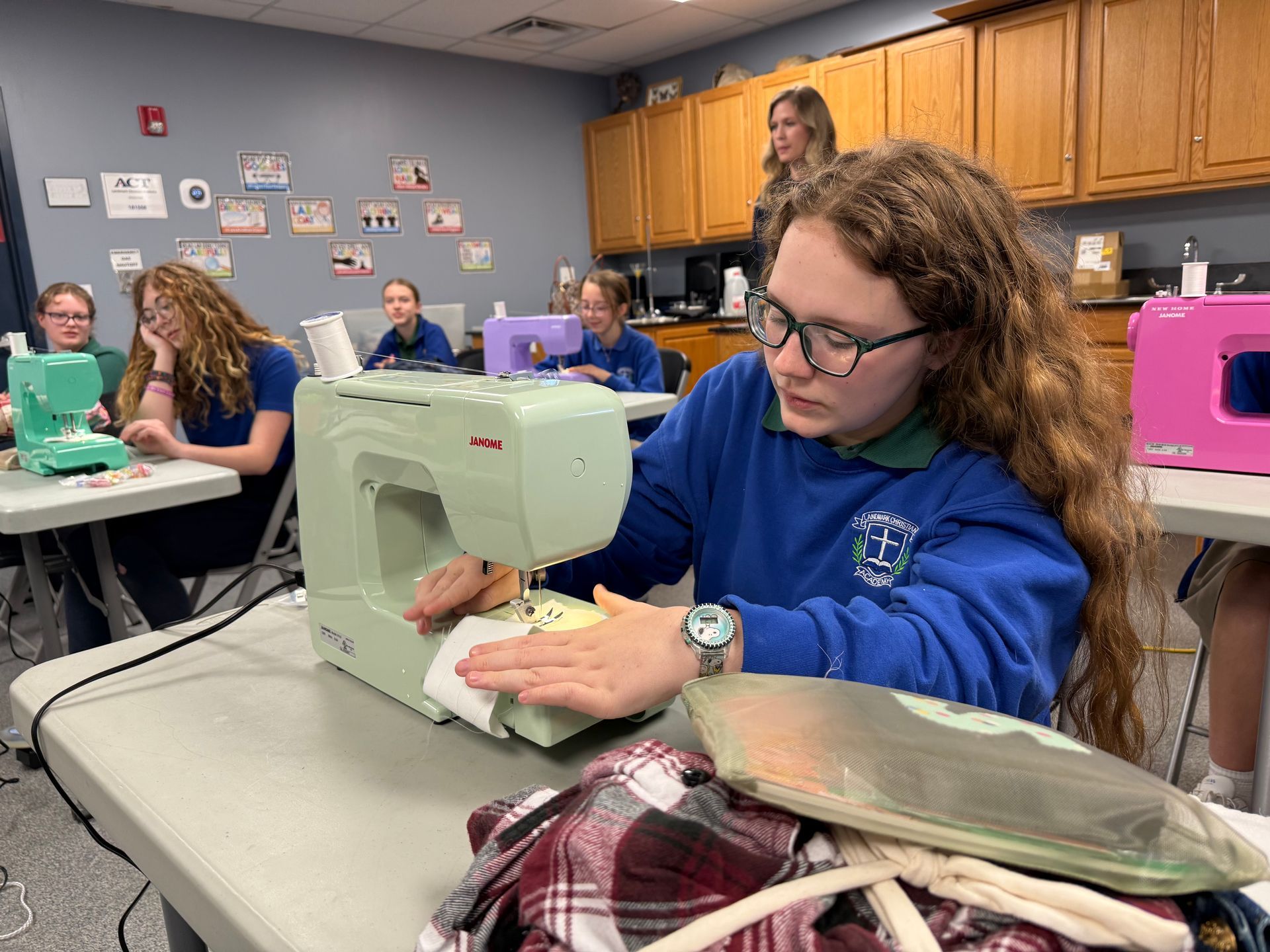 Student using a sewing machine in a classroom workshop, with others working at tables nearby