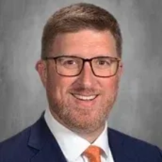 Professional headshot of a smiling man wearing glasses, a navy suit, and an orange tie against a gray background