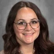 Portrait of a smiling person with glasses and dark wavy hair against a gray background