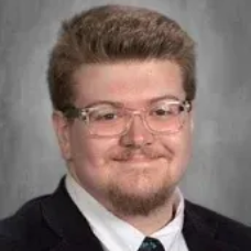 Portrait of a man in a suit and glasses, smiling against a gray studio backdrop
