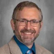 Headshot of a smiling man with glasses, beard, tan jacket, and blue shirt against a gray background