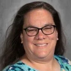 Smiling person with glasses and long dark hair in a blue patterned top against a gray background