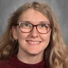 Portrait of a smiling person with glasses and wavy blonde hair against a gray background