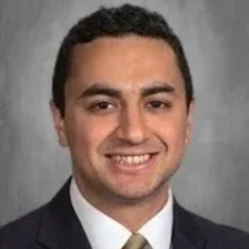 Studio headshot of a smiling man in a dark suit, white shirt, and tie against a gray background