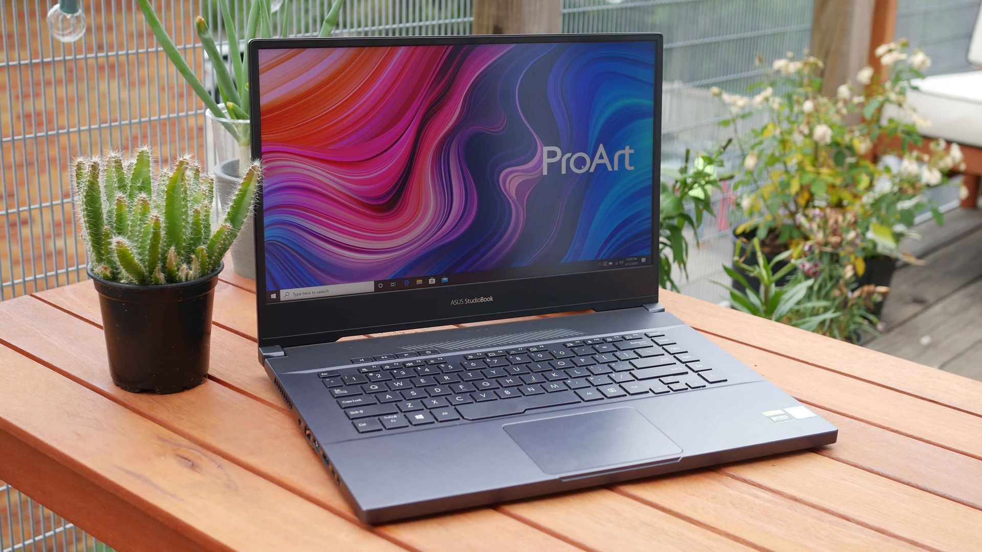Laptop on a wooden patio table beside a potted plant, with a colorful ProArt screen.