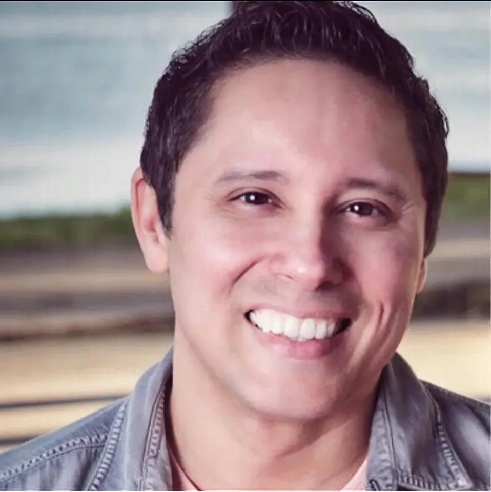 Man smiling at the camera. He has dark hair and is wearing a grey jacket. Background is blurred ocean view.