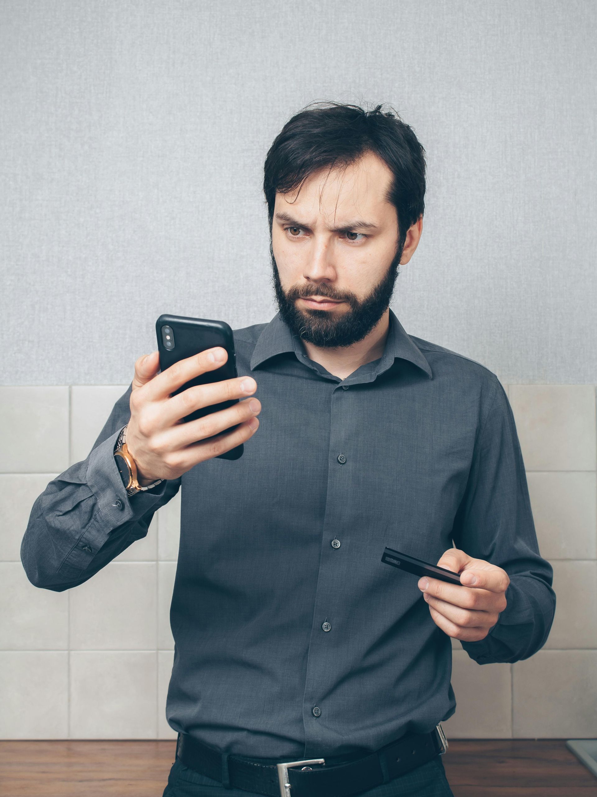 A man with a beard is holding a credit card and looking at his phone