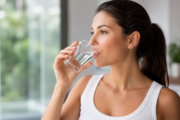 Una donna con una canottiera bianca beve acqua vicino a una finestra.