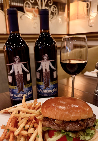 Two wine bottles, a glass of red wine, a burger with fries on a plate.