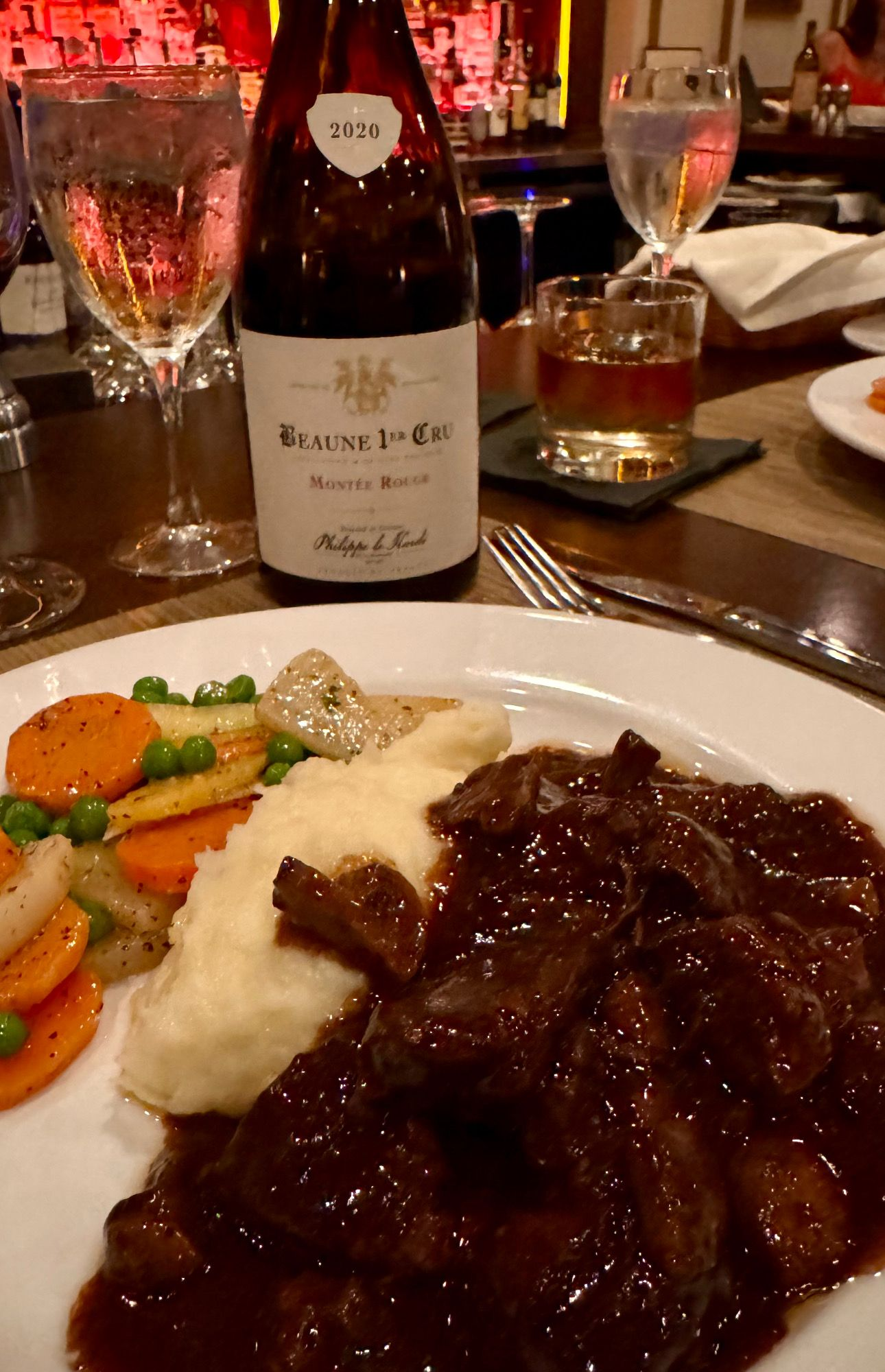 Plate of meat with gravy, mashed potatoes, and vegetables, wine bottle, glasses, and a cocktail on a table.