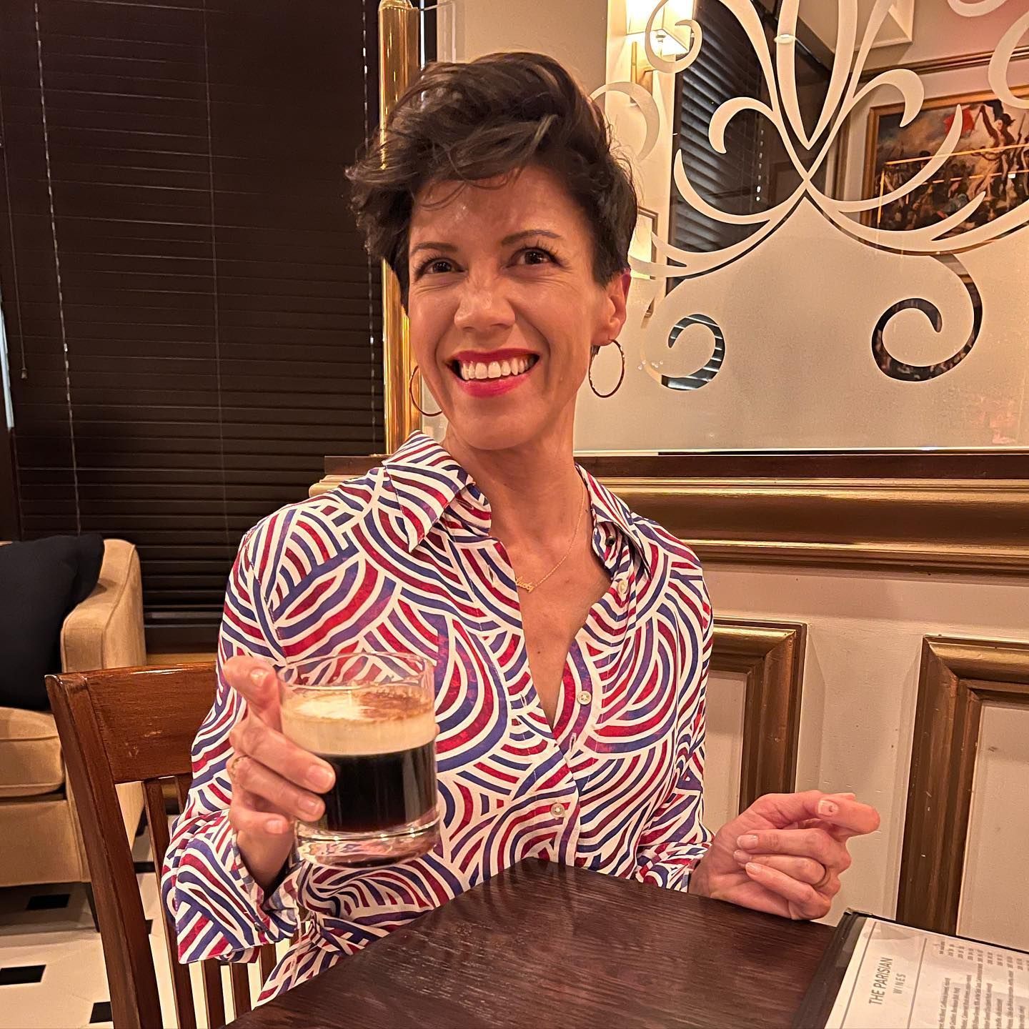 Woman smiling, holding a dark drink, wearing a patterned shirt, inside a restaurant.