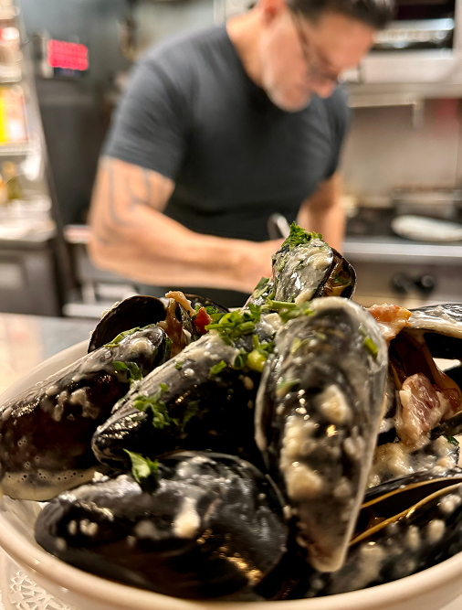 Bowl of steamed mussels with sauce; chef blurred in background.