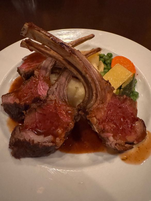 Rack of lamb with sauce, mashed potatoes, peas, and carrots on a white plate.