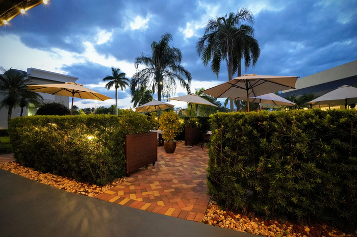 Outdoor restaurant patio with umbrellas, hedges, and palm trees under a cloudy sky.