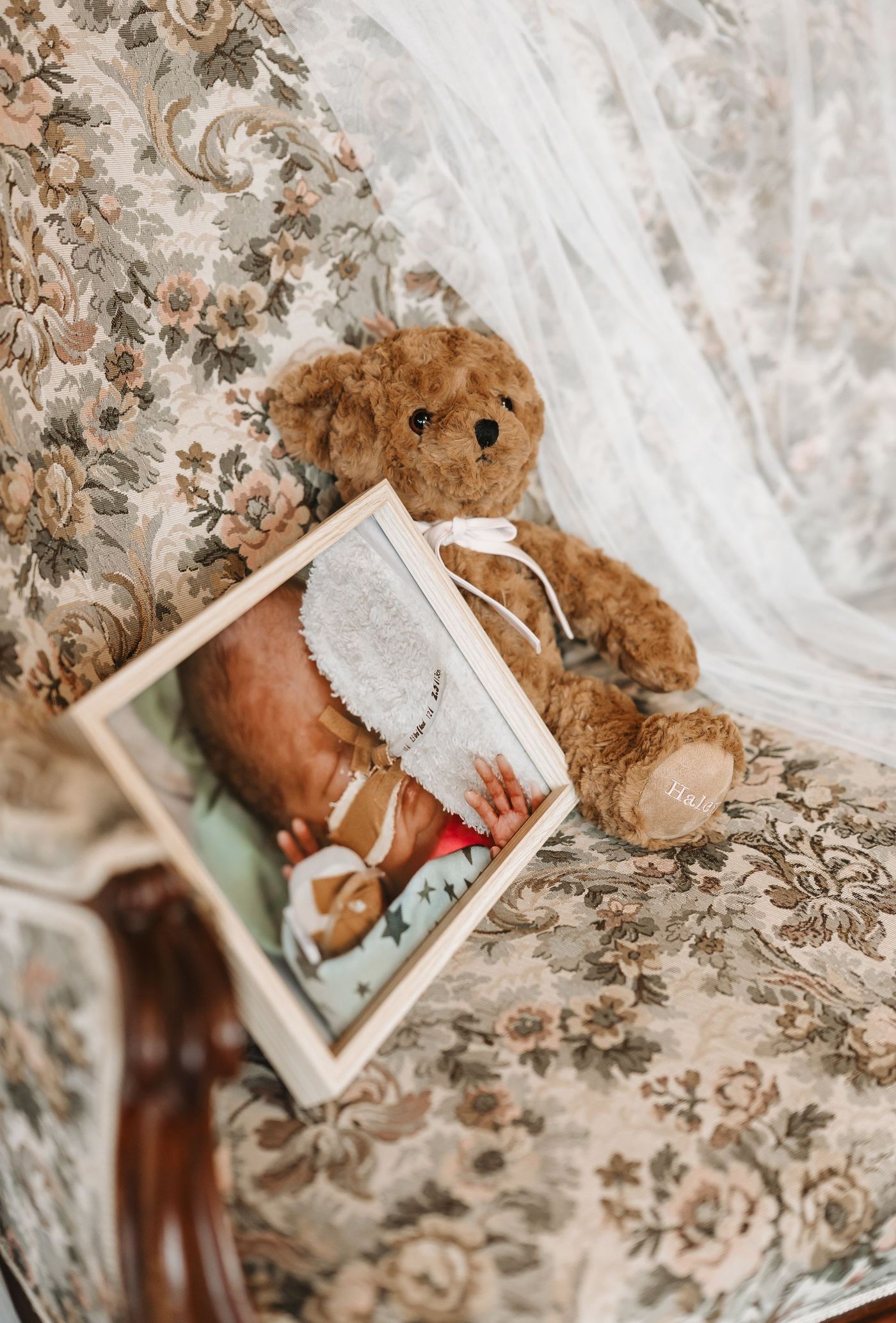 Een teddybeer naast een ingelijste foto van een pasgeborene op een bank met bloemenprint, met een doorschijnend wit gordijn er vlakbij.