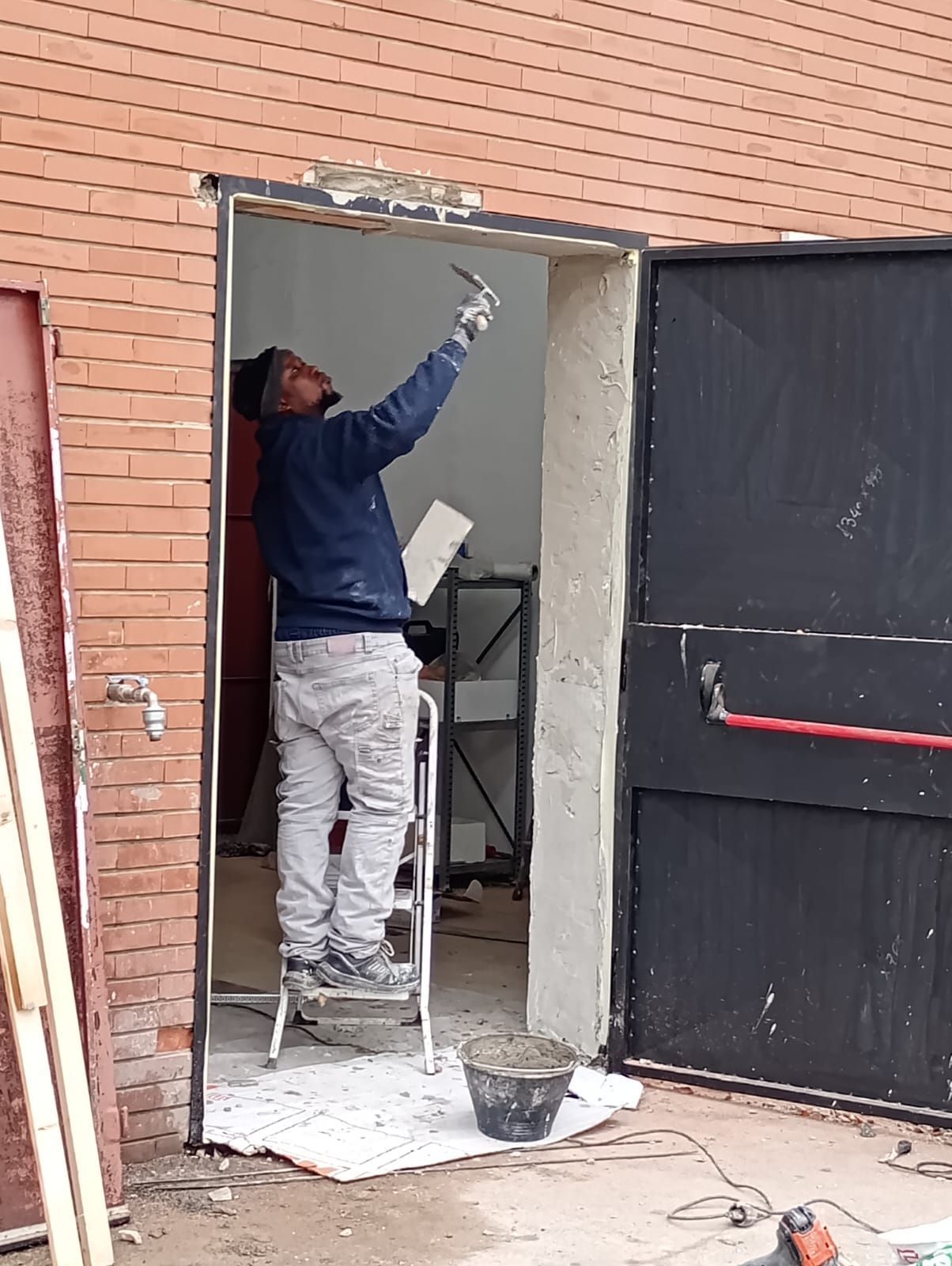 Uomo su una scala che applica l'intonaco attorno allo stipite di una porta. Edificio in mattoni, porta nera, secchio e attrezzi visibili.