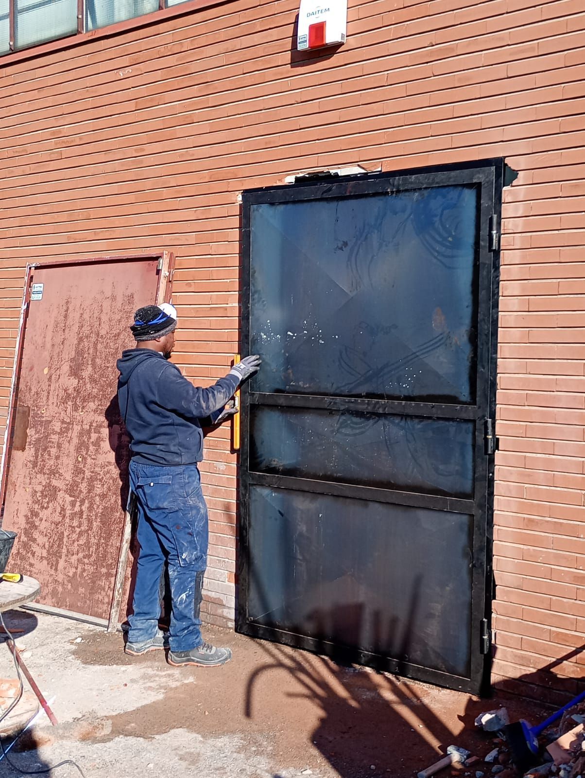 Persona in abiti da lavoro che installa una grande porta di metallo nero sulla facciata di un edificio in mattoni.