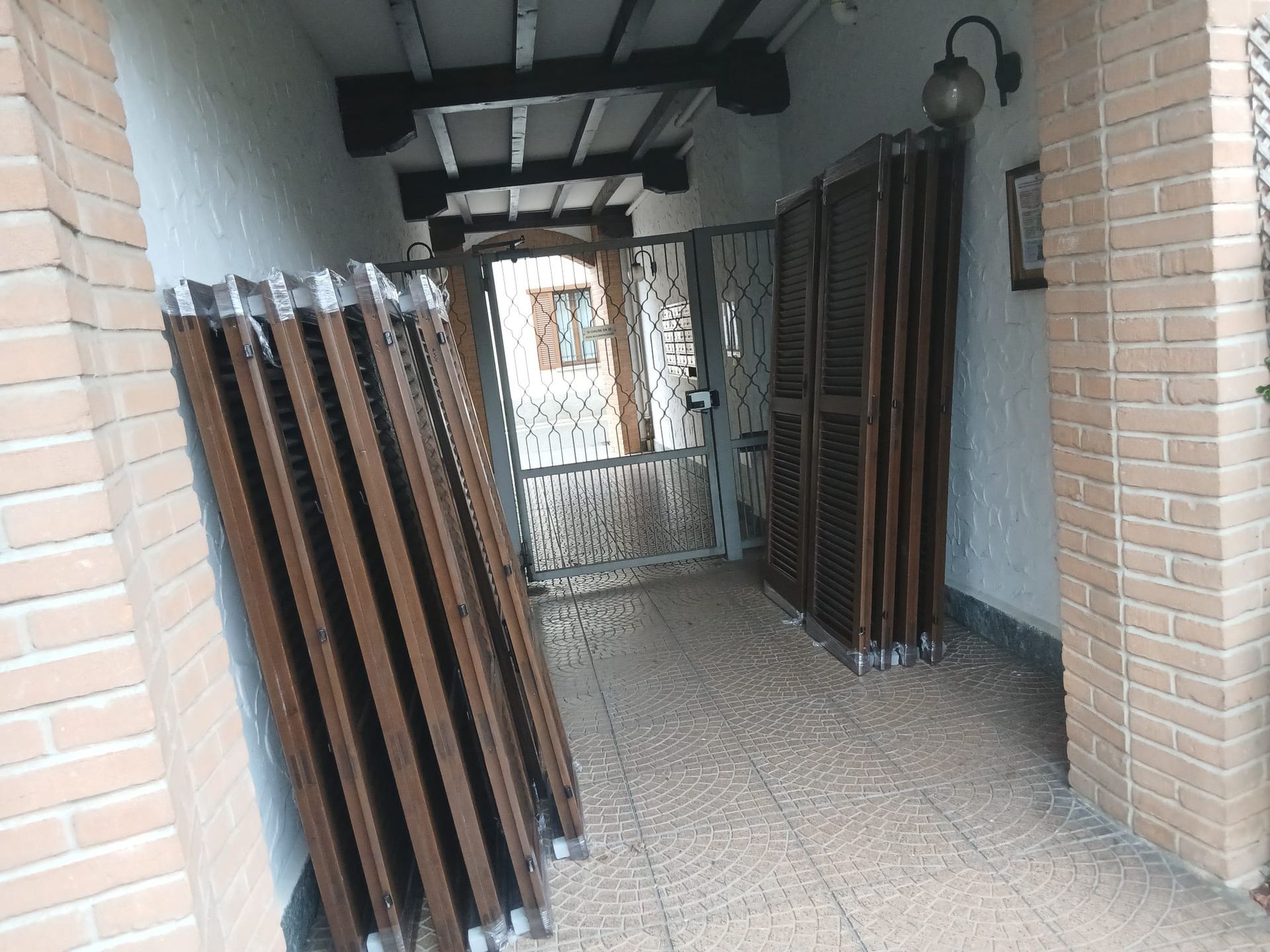 Ingresso interno con persiane a soffietto marroni ripiegate e chiuse, che conduce a un cancello e a una porta.