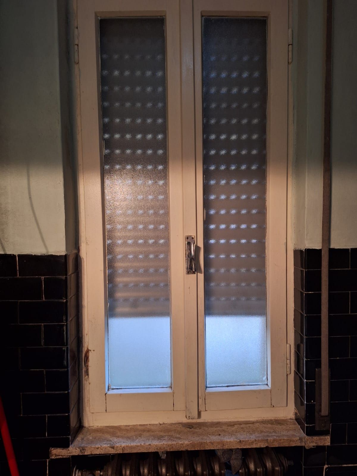 Finestra con cornice bianca, vetro opaco e tenda avvolgibile chiusa. Situata in una stanza con piastrelle scure e un termosifone sottostante.