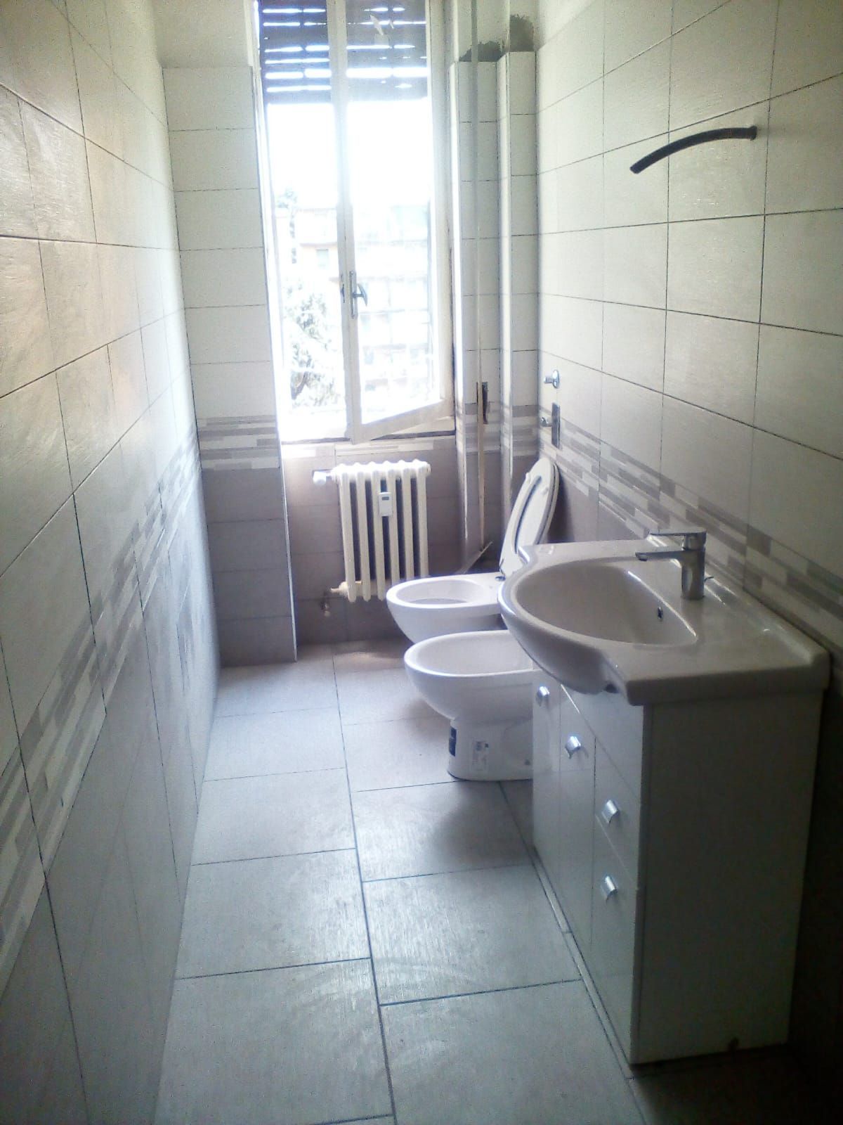 Bagno con lavandino, WC, bidet e finestra. Pareti piastrellate bianche e grigie.