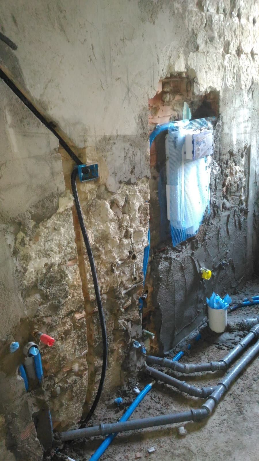 Lavori idraulici in corso: tubi blu, tubi grigi, muro di mattoni a vista, fili neri, costruzione incompiuta.