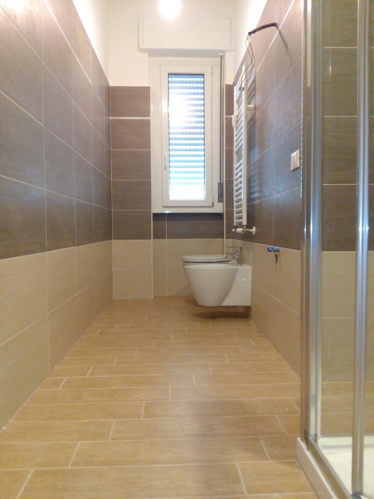 Bagno con pareti piastrellate beige e marroni, WC, finestra con persiane e box doccia.