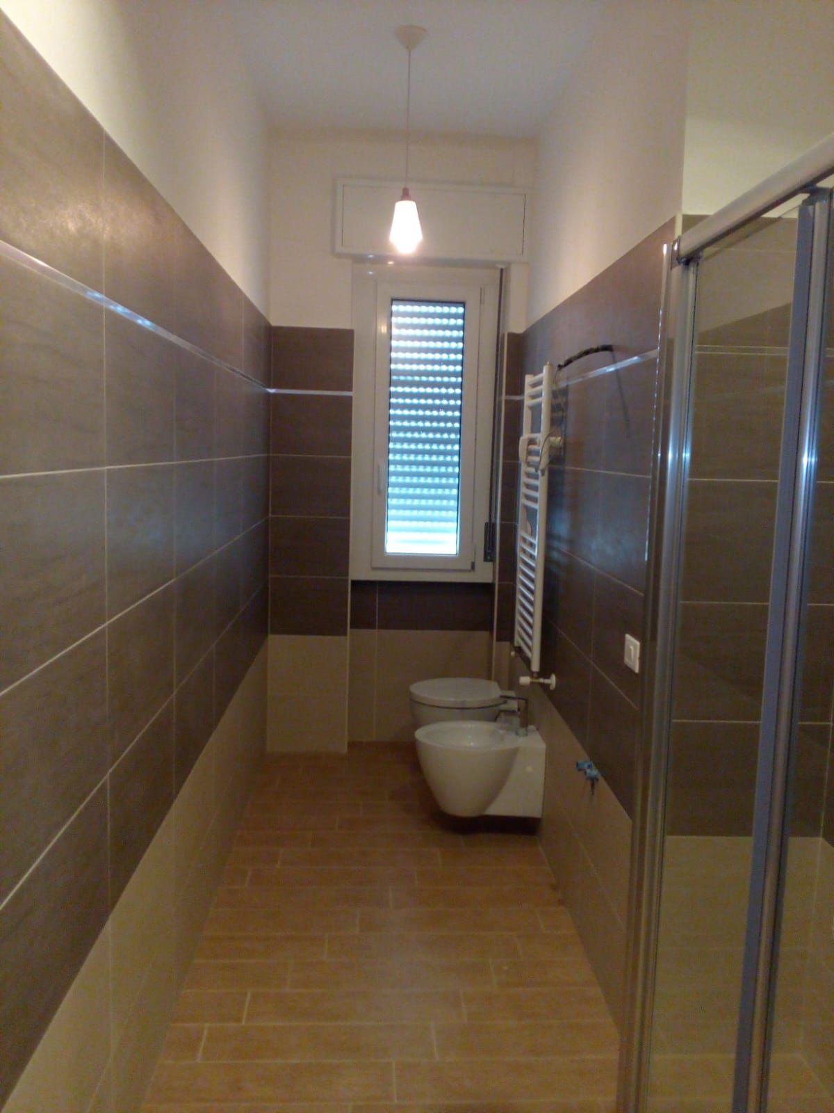 Bagno stretto con piastrelle color sabbia e pareti in piastrelle marroni, finestra, WC, bidet, box doccia e scaldasalviette.