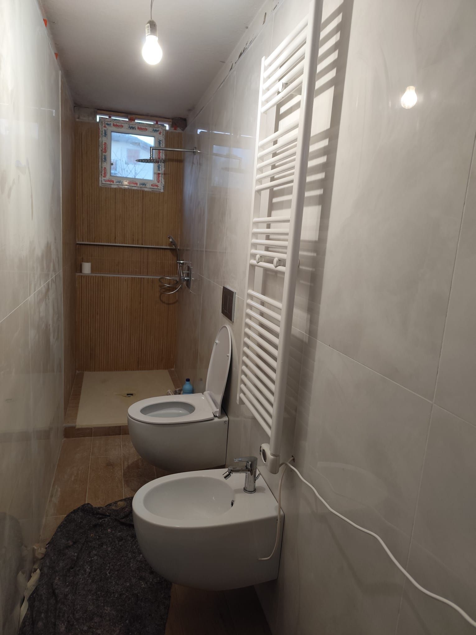 Bagno con WC, bidet, scaldasalviette, doccia e finestra; pareti piastrellate grigio chiaro e pavimento piastrellato beige.