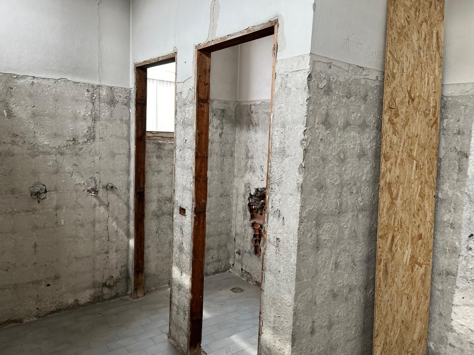 Vista interna di un bagno in costruzione con due porte, pareti in cemento e un'asse di legno.
