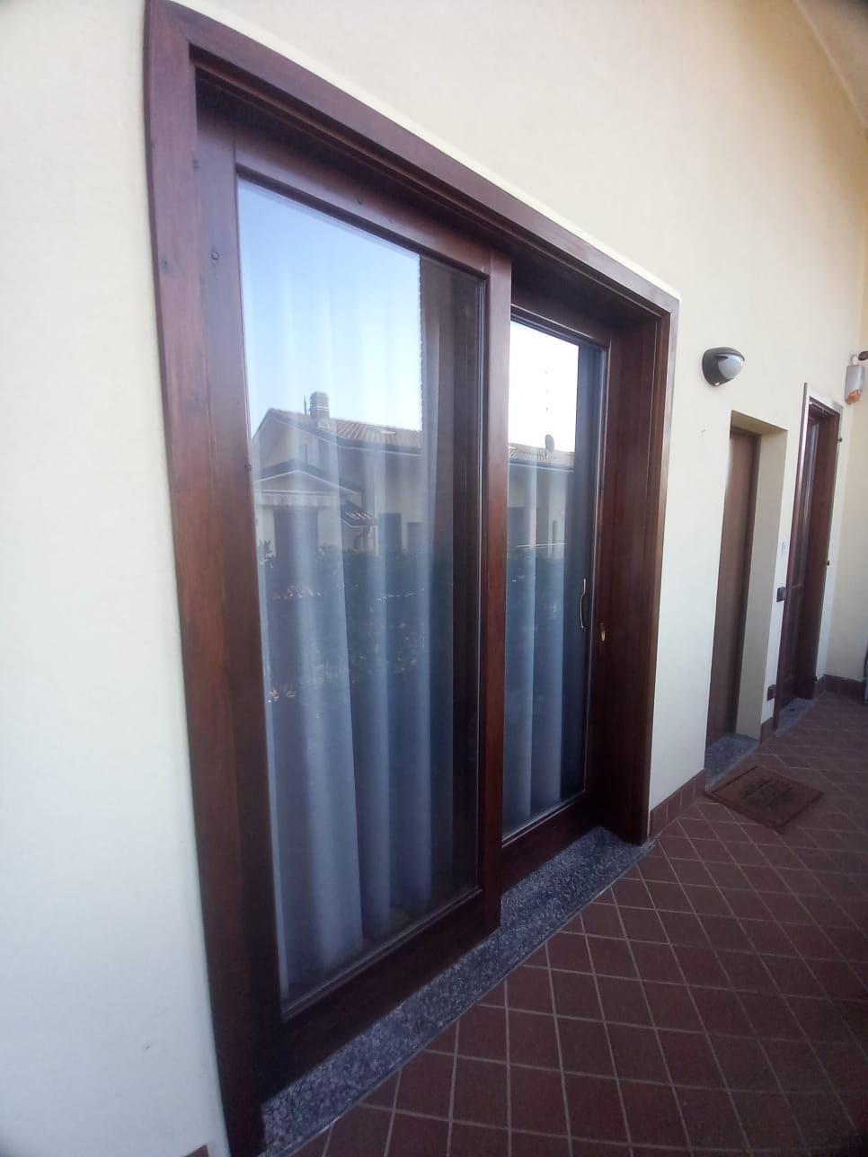 Porte scorrevoli in vetro con telaio in legno marrone scuro, edifici riflettenti e tende bianche, su un patio in mattoni.