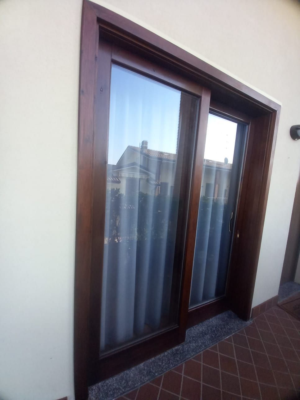 Porta scorrevole in vetro con telaio in legno su una parete color cuoio. Telaio della porta marrone, riflesso del cielo azzurro, tende bianche all'interno.