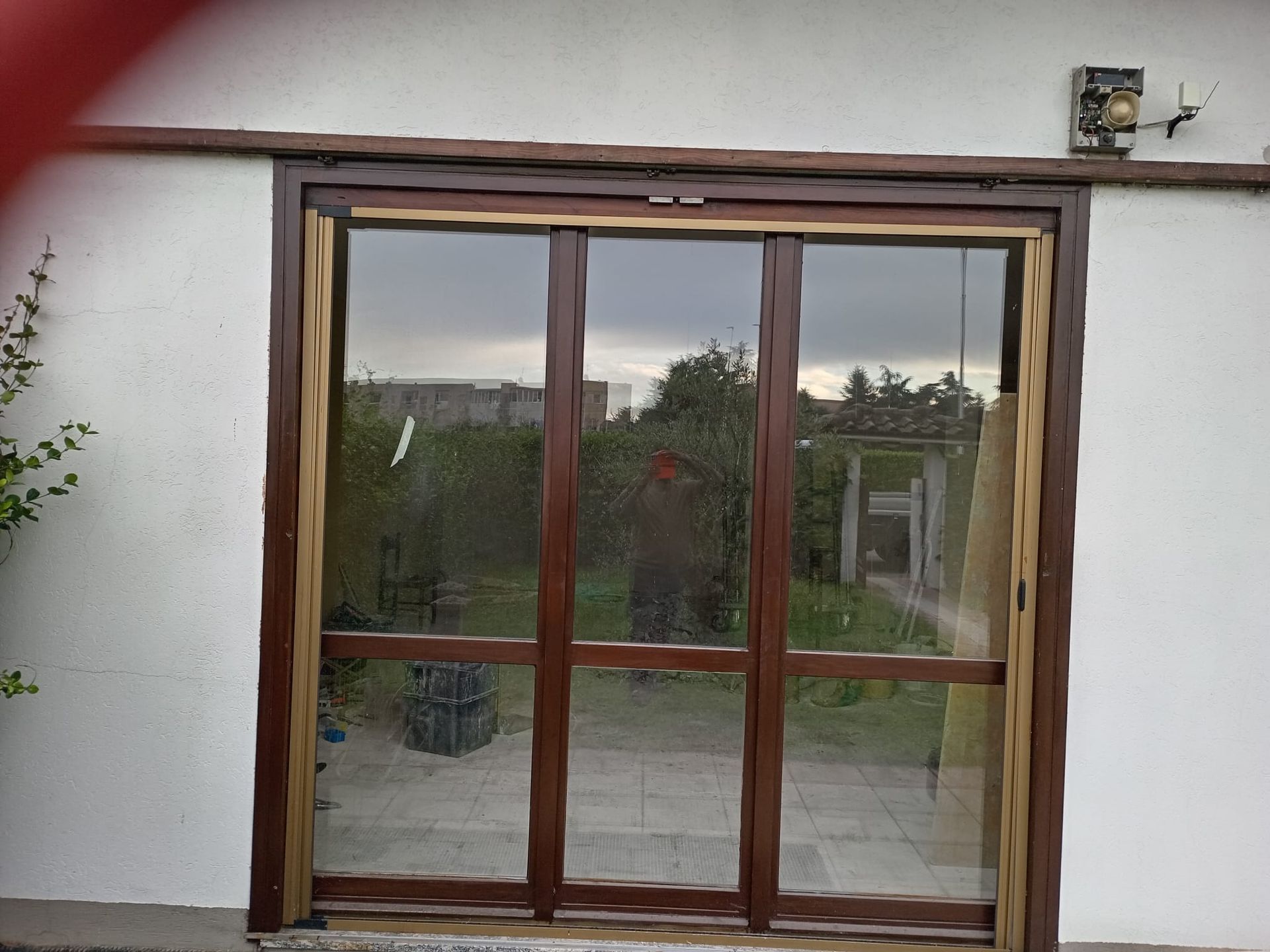 Porte scorrevoli in vetro con telaio marrone e zanzariere, che si aprono su un cortile con alberi e un cielo nuvoloso.