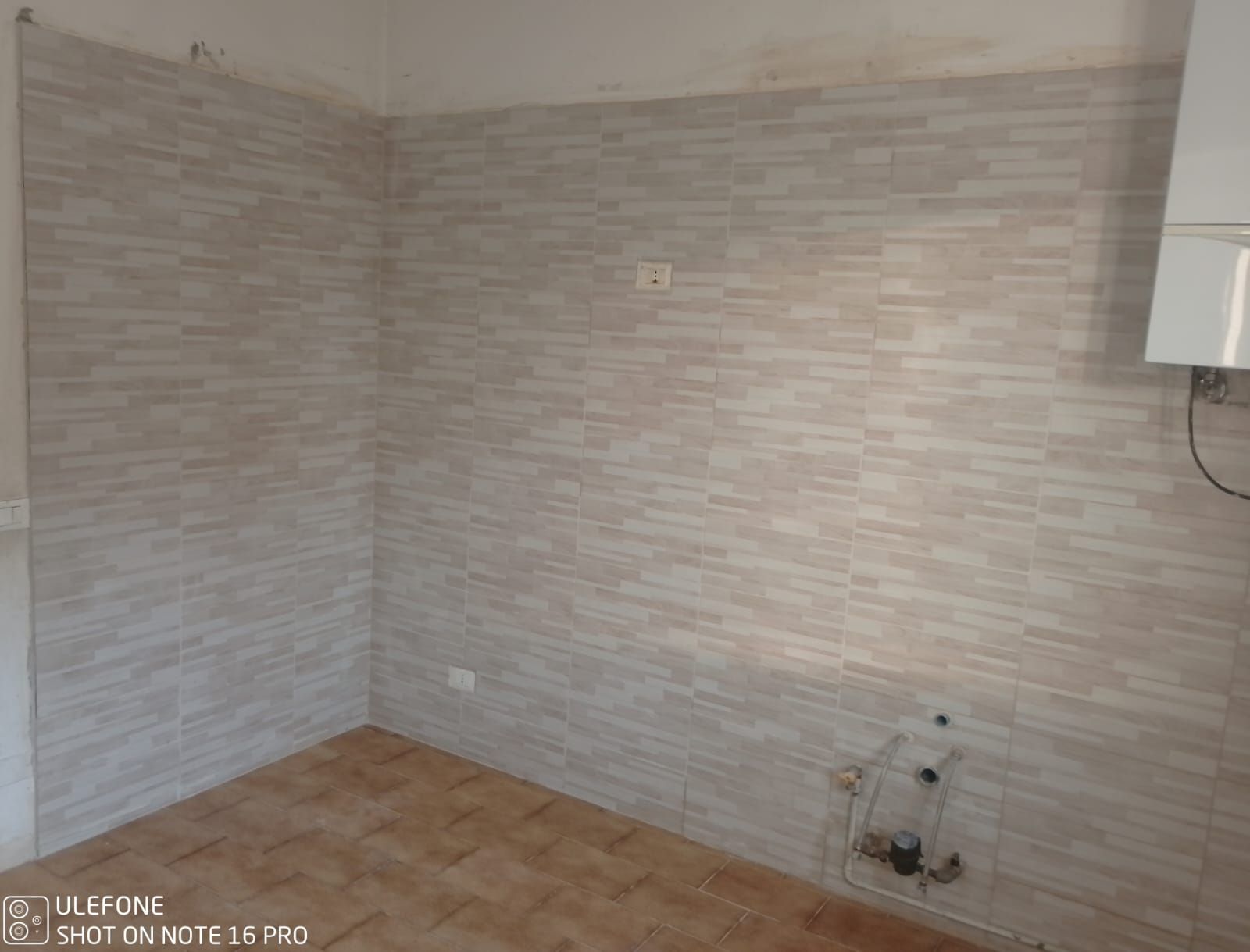 Angolo cottura vuoto con pareti e pavimento piastrellati, tubi e scaldabagno.