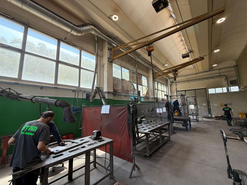 Operai in una grande officina che azionano macchinari vicino a tavoli e attrezzature metalliche.