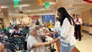 A person in a yellow outfit performs for a group of seniors sitting in wheelchairs in a decorated indoor activity room.