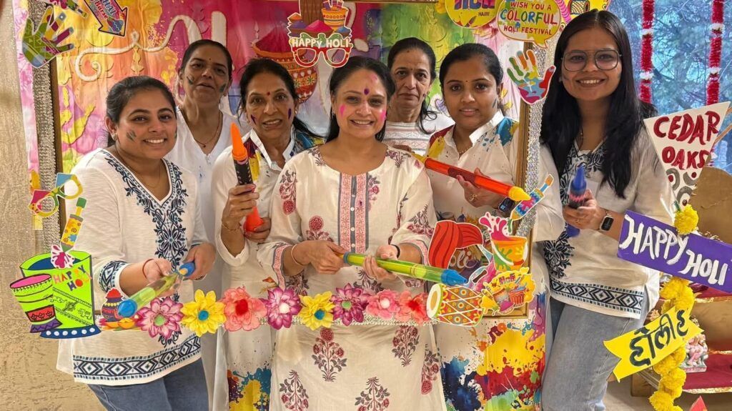Six people smiling and posing together behind a colorful, decorated Holi festival photo frame indoors.